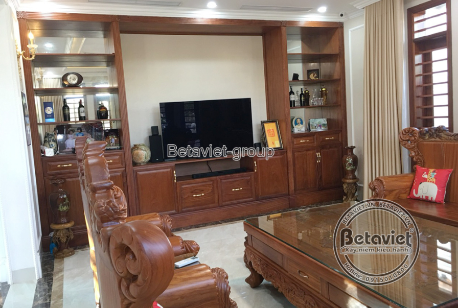 Góc phòng khách nhìn từ phía đối diện, tập trung vào hệ tủ kệ TV lớn bằng gỗ tự nhiên màu nâu trầm, tích hợp nhiều ngăn ch…