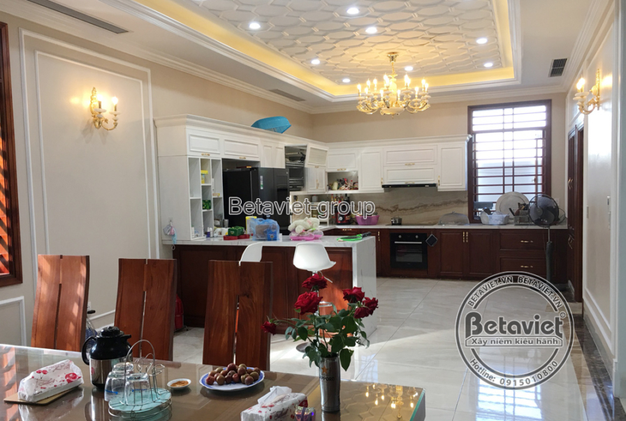 Góc nhìn tổng thể không gian bếp và phòng ăn liên thông, nổi bật với tủ bếp chữ L màu trắng kem và nâu gỗ, mặt bếp ốp đá…