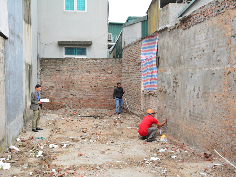 Góc nhìn khác của buổi khảo sát mặt bằng thi công, tường gạch bao quanh khu đất trống chưa xây. Người mặc sơ mi tay cầm gi…