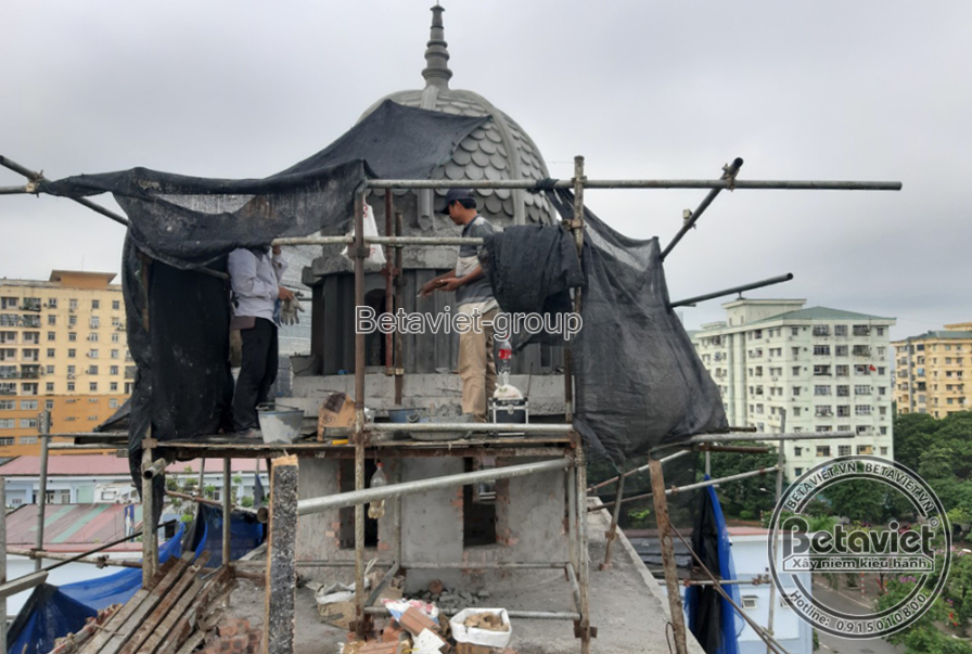 Khối tháp mái vòm được che bằng lưới đen tạm, chóp nhọn nhô cao giữa các tòa nhà cao tầng phía xa. Hai công nhân đứng trên…