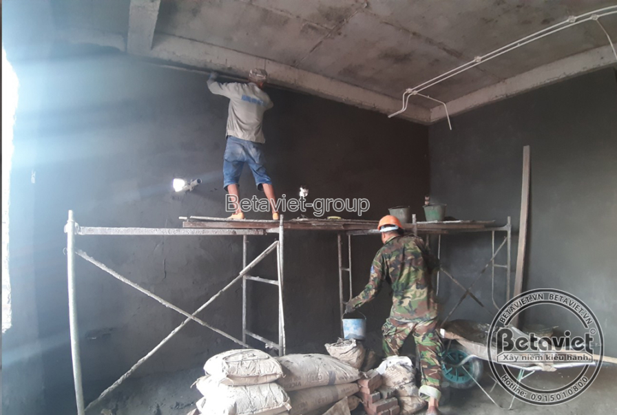 Không gian phòng bên trong còn thô mộc với tường và trần trát vữa xám, ống điện âm tường chạy dọc góc trần. Một công nhân…