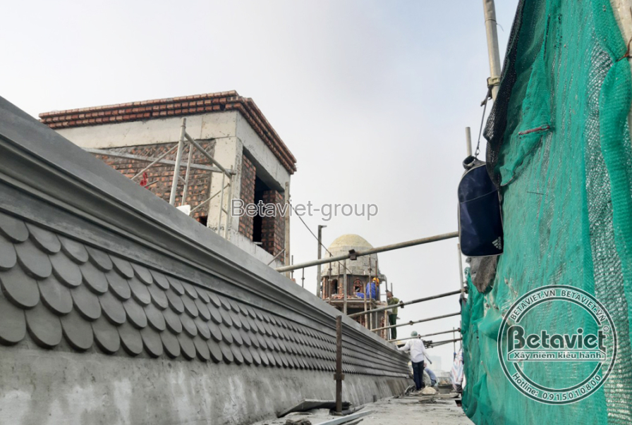 Góc nhìn từ trên cao dọc theo phần tường chớp mái có đường viền ngói vảy cá phủ kín, lưới xanh phủ tạm phía trên. Một tháp…