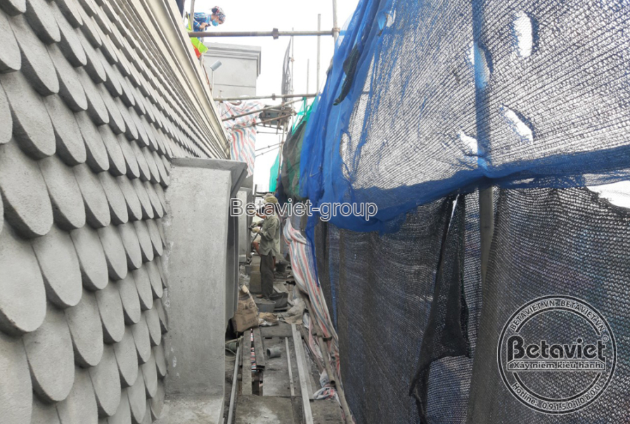 Cận cảnh bề mặt ngói vảy cá màu xám ốp trên mái vòm, lưới bảo hộ xanh và bạt che phủ giàn giáo phía trên. Từng viên ngói đ…