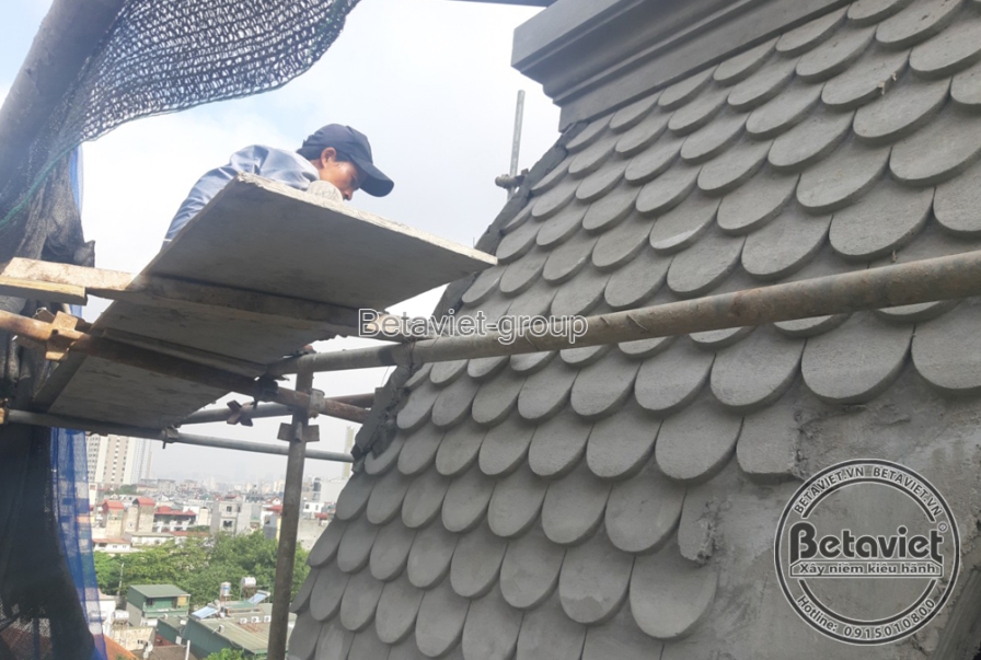 Công nhân đang thi công ốp mái vảy cá màu xám cho phần mái vòm của biệt thự, sử dụng giàn giáo và lưới bảo hộ màu xanh. Gó…