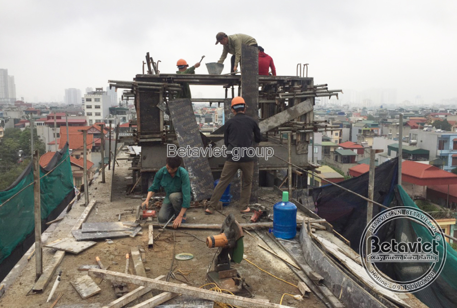 Công nhân trên sân thượng đổ bê tông vào khung ván cong bằng xô chuyền tay, cọc giàn giáo bao quanh, bình gas nhỏ đặt gần…