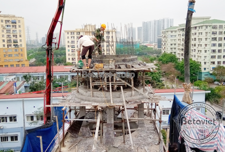 Toàn cảnh khối tháp mái vòm tròn đang đổ bê tông, cần bơm bê tông vươn tới đỉnh tháp, công nhân đứng trên giàn giáo thao t…