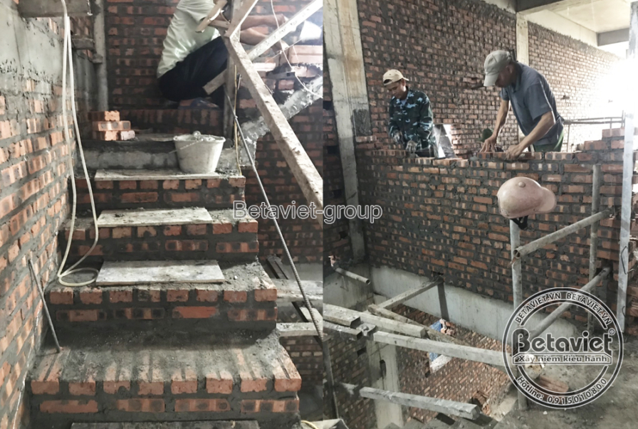 Bức tường gạch đỏ cạnh khung cầu thang gỗ tạm, một công nhân leo lên khung như bậc thang, người còn lại xây gạch trên bục…