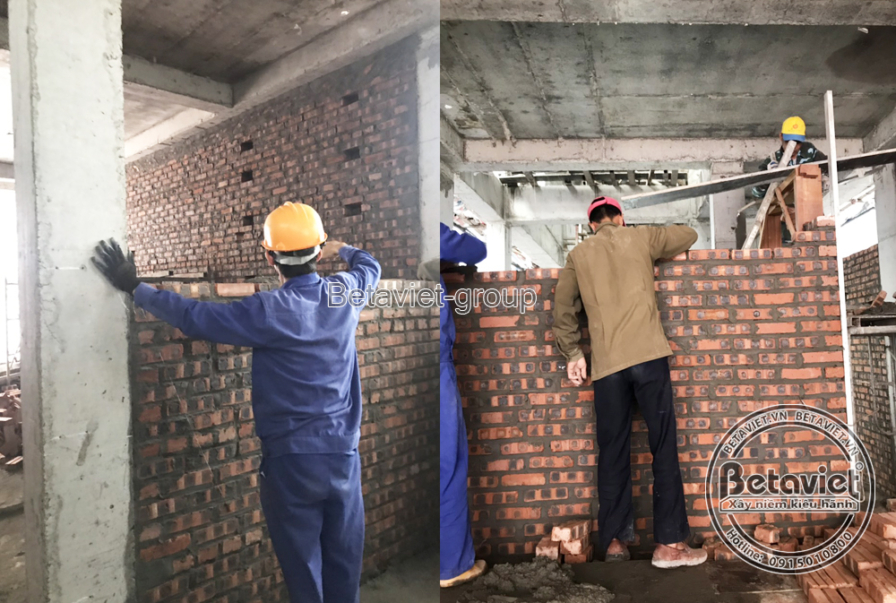 Hai công nhân xây tường gạch đỏ nối giữa cột và dầm bê tông lộ thiên, không gian sàn còn mở thông ra ngoài. Ánh sáng tự nh…