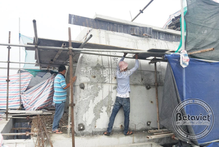 Hai công nhân thao tác cạnh bức tường bê tông cốp pha còn dấu ván khuôn, một người đẩy cây chống giàn giáo sát tường. Bạt…