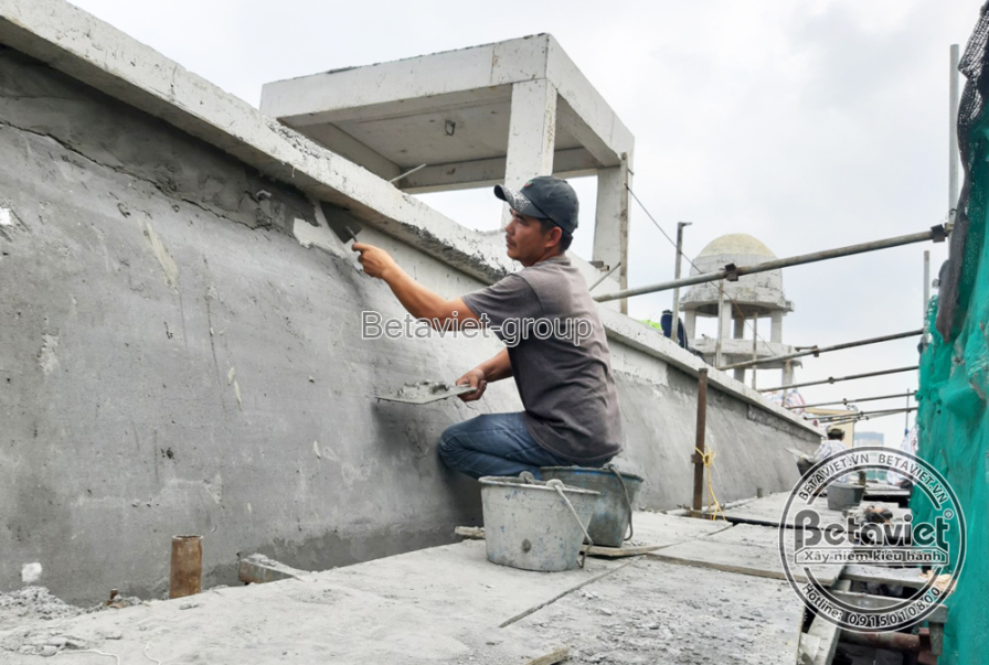 Công nhân quỳ trên mép sân thượng trát vữa hoàn thiện bề mặt tường lan can, phía sau là khối mái vòm nhỏ. Xô vữa đặt cạnh…