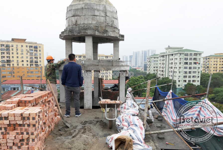 Khối tháp mái vòm có cột đỡ dạng đình bát giác trên nóc tòa nhà, hai công nhân đứng cạnh chồng gạch nhìn ra toàn cảnh thàn…