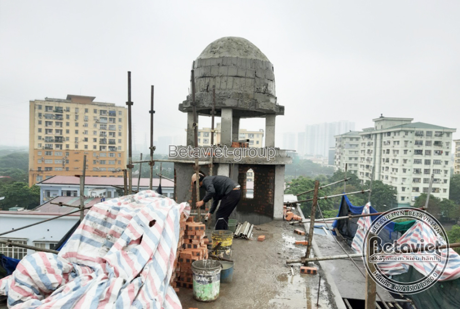 Khối tháp mái vòm nhìn từ sân thượng, một công nhân xây gạch tại chân tháp, vật liệu bạt sọc chất đống gần đó. Phía xa là…