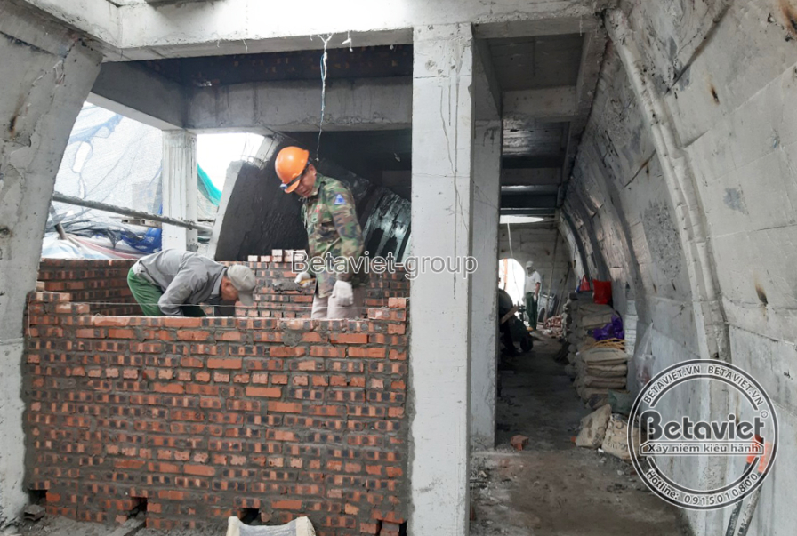Công nhân đội mũ bảo hộ xây tường gạch đỏ giữa hai cột bê tông, không gian sàn tầng mở rộng đón ánh sáng tự nhiên từ mặt t…