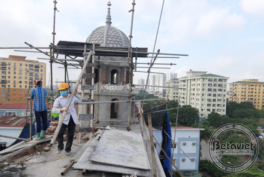 Đỉnh tháp mái vòm đang lắp phần chóp, hai công nhân đứng trên giàn giáo cao thao tác giữa nền trời cùng các khối nhà cao t…