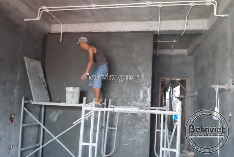 Góc phòng trát vữa hoàn thiện với ống điện âm tường chạy dọc trần, một công nhân đứng trên giàn giáo nhỏ vươn tay xử lý gó…