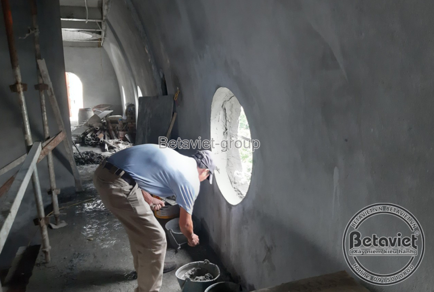 Tường cong nội thất với ô cửa sổ tròn, một công nhân cúi người láng vữa mép sàn sát chân tường. Bề mặt trát nhẵn màu xám…