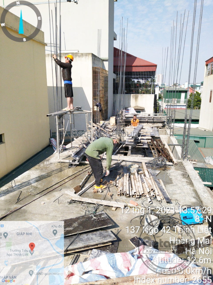 Công nhân đứng trên giàn giáo buộc lồng thép cột theo phương thẳng đứng, sát bên là sàn lưới thép đang chờ đổ bê tông. Bức…