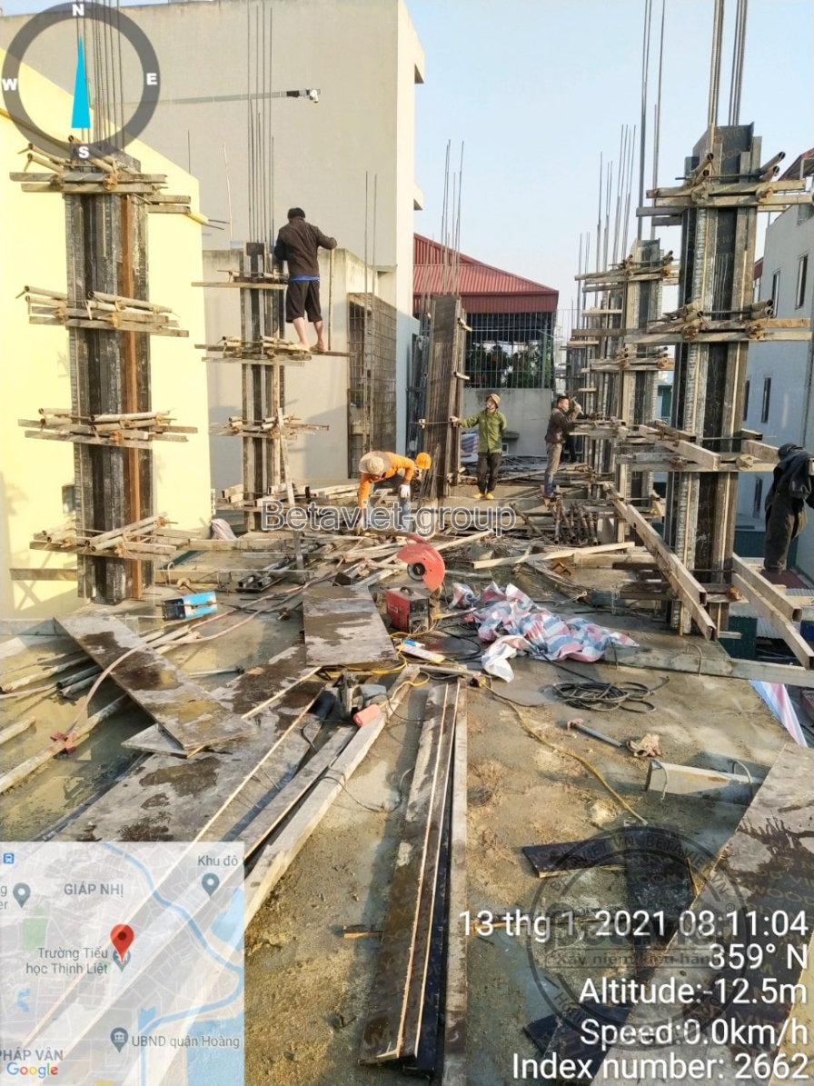 Hệ cột thép chờ dựng thẳng đứng từ sàn bê tông, khung gỗ cốp pha bao quanh chân cột. Nhiều công nhân rải rác làm việc trên…