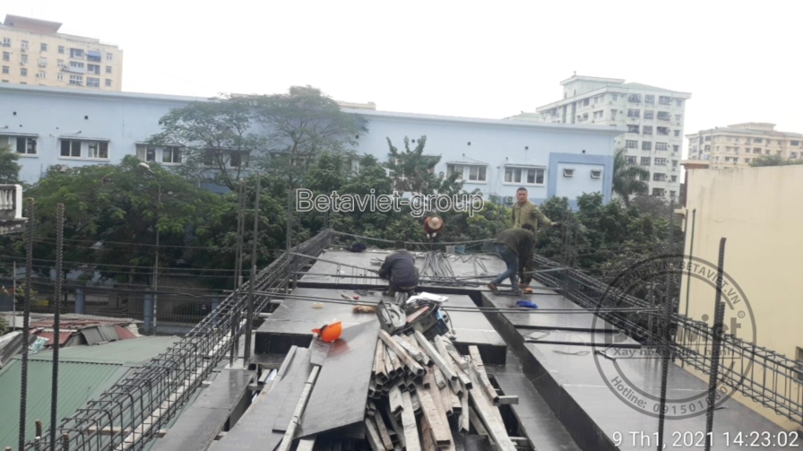 Sàn lưới thép hoàn tất chờ đổ bê tông, gỗ ván khuôn thừa chất đống ở mép sàn. Hai công nhân đứng phía xa trò chuyện, nhà d…