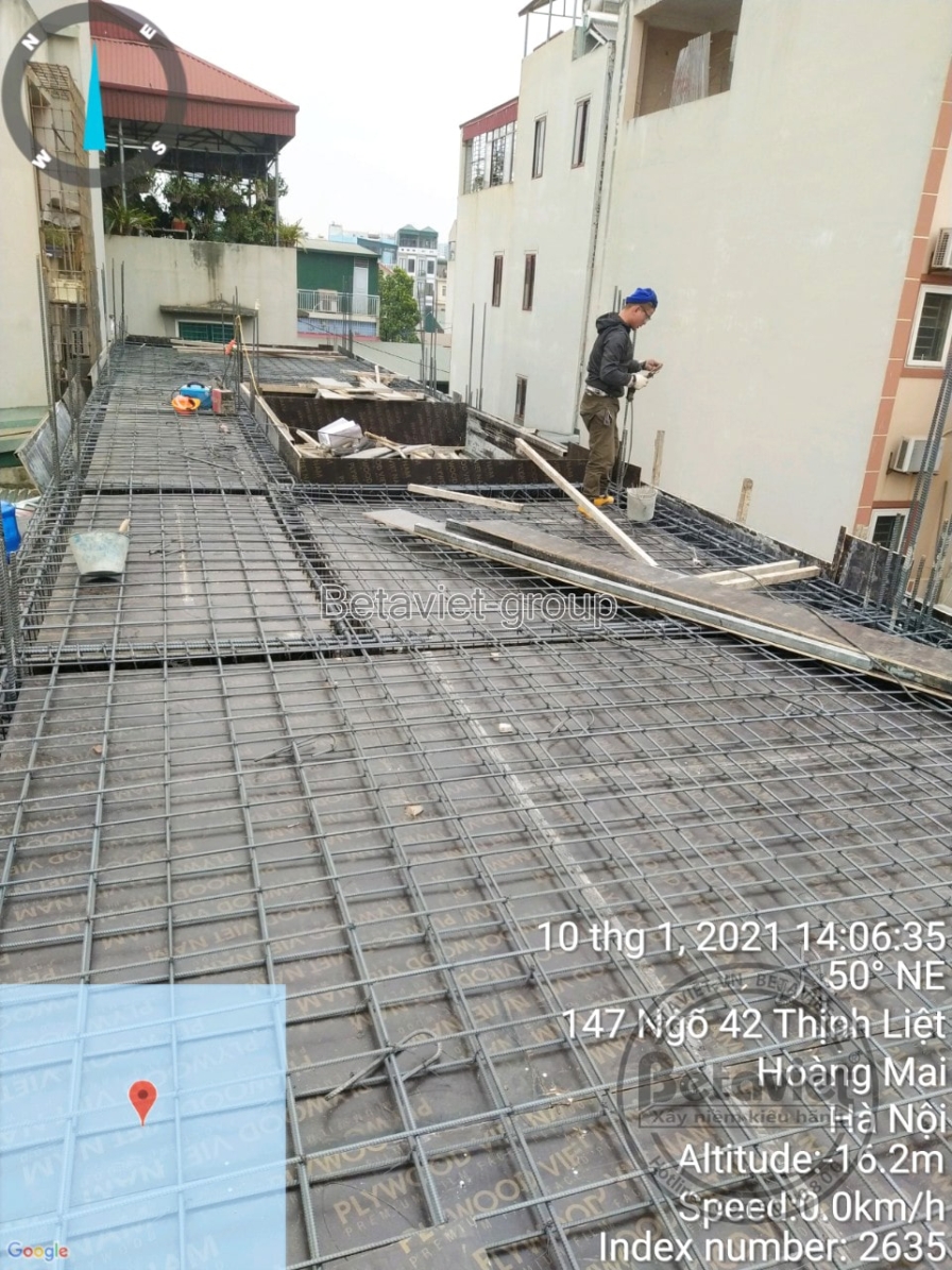 Sàn mái đang đổ bê tông với lưới thép sàn đan kín, một công nhân đứng ở góc xa kiểm tra. Xung quanh là các mái nhà dân cư…