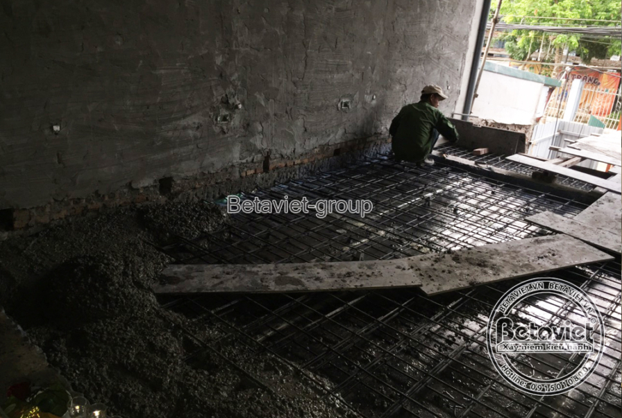 Căn phòng còn thô ráp với tường bê tông chưa trát, một công nhân cúi láng vữa nền nhà bằng bay. Ánh sáng tự nhiên hắt vào…