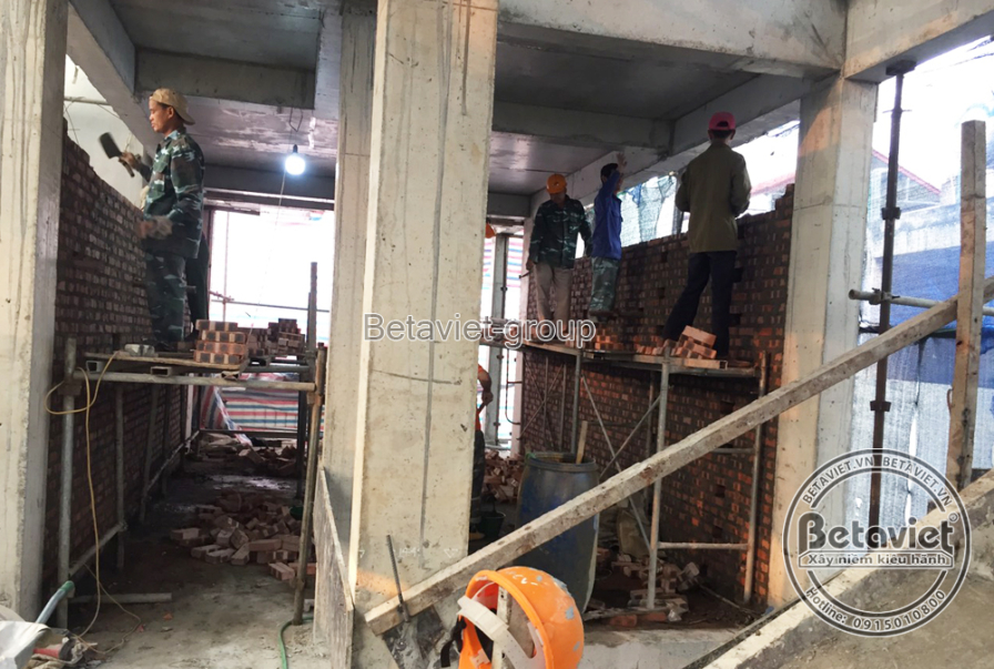 Sàn tầng đang xây với hệ cột bê tông và mép sàn lộ thiên, công nhân xây tường gạch đỏ giữa các cột, một người đứng trên vá…