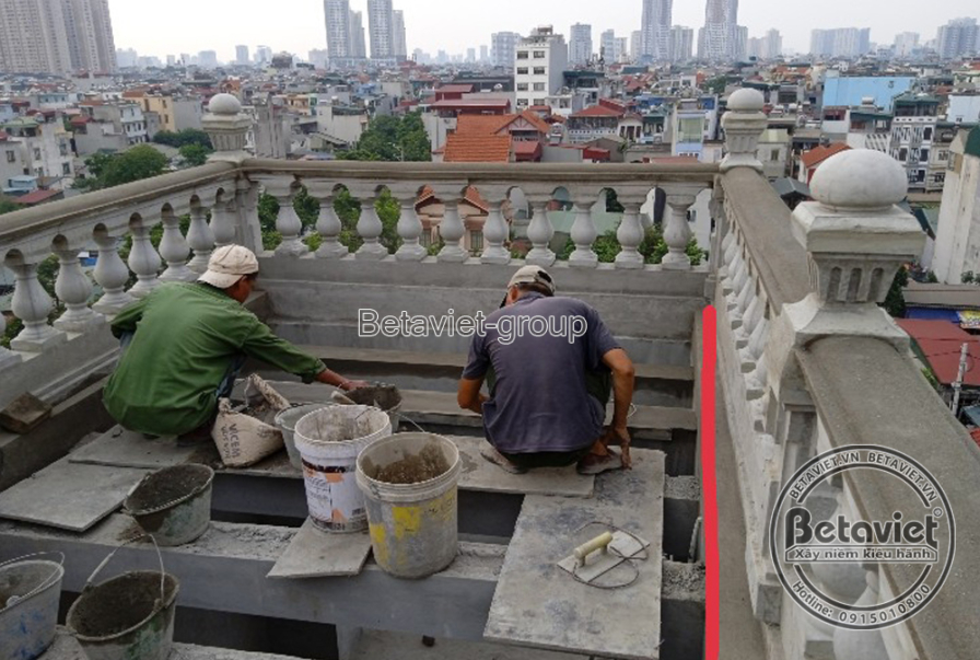 Không gian sân thượng nhìn qua hàng lan can con tiện đã đúc hoàn chỉnh, một công nhân quỳ trát chân lan can. Phía xa là to…
