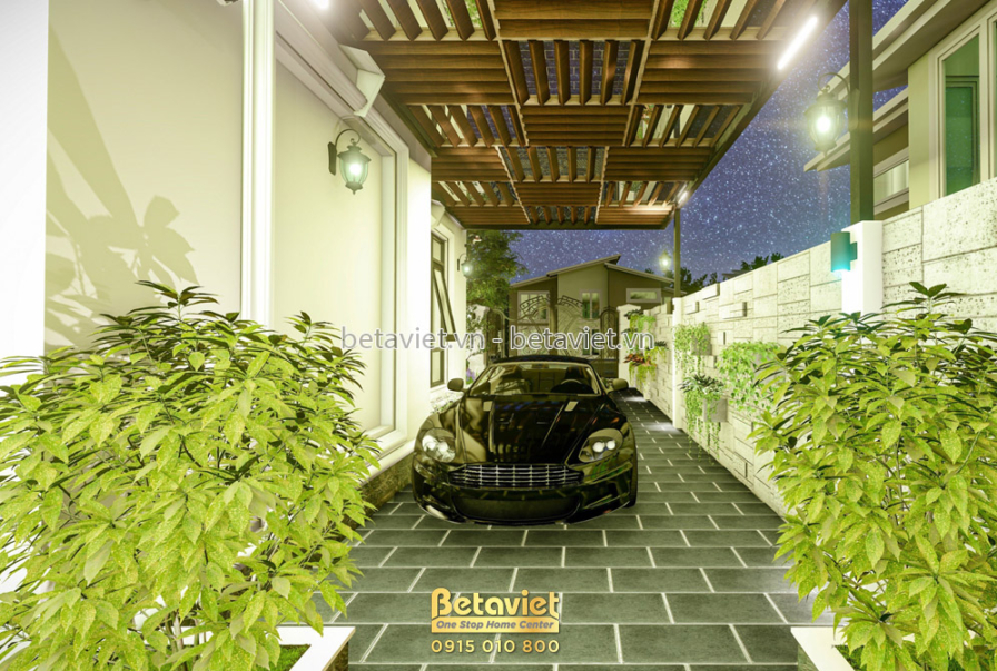 Hành lang để xe có mái pergola gỗ dạng nan dài, xe màu tối đậu giữa hai hàng cây bụi rậm rạp, nền lát đá vuông sáng màu, k…