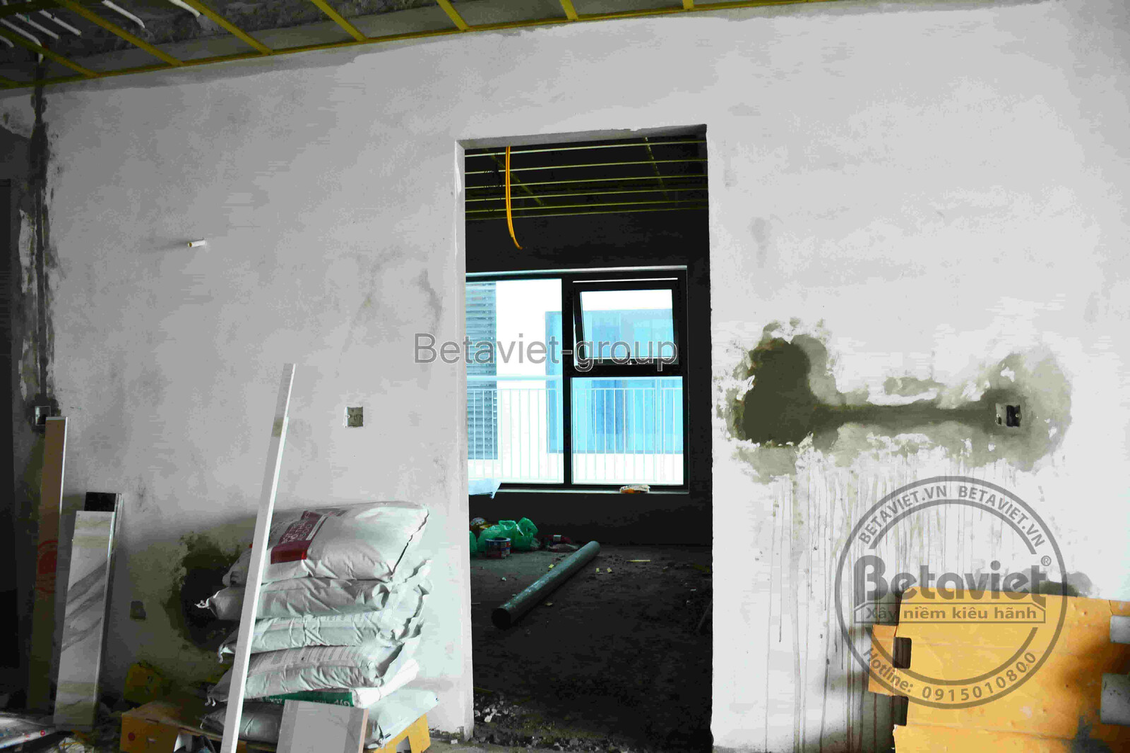 Góc tường trát vữa xám dở dang cạnh lối cửa đi thông phòng, bao vật liệu xây dựng xếp gọn, view cửa kính phía sau. Không g…