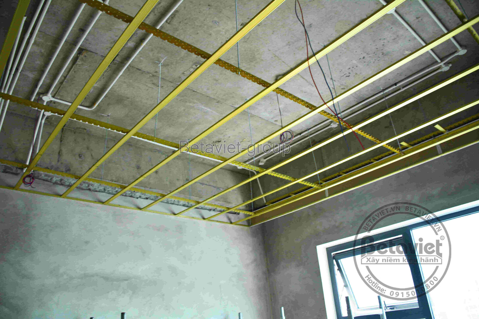 Trần nhà đang lắp khung xương thép vàng cho hệ trần thạch cao, tường bê tông thô, cửa kính lớn phía dưới đón ánh sáng tự n…