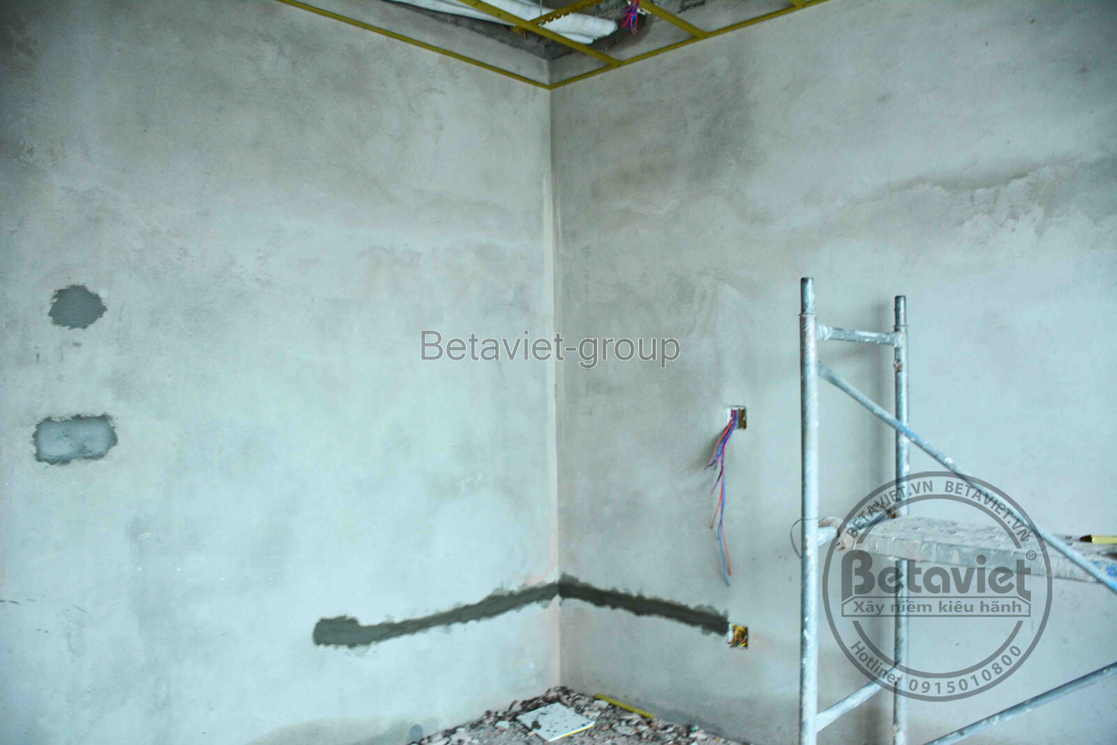 Góc tường phần thô với các mảng vá vữa xám đậm xen kẽ, dây điện chờ nối, khung nhôm dựng tạm ở góc phòng. Trần bê tông thô…