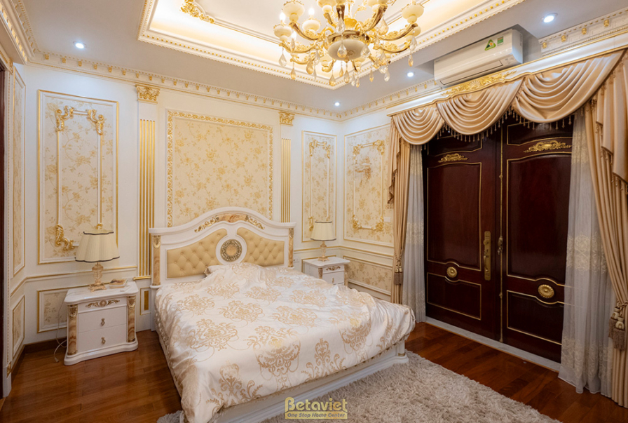 Phòng ngủ với giường lớn, tủ quần áo gỗ tự nhiên màu nâu đỏ. Tường ốp phào chỉ và giấy dán tường. Rèm cửa hai lớp màu kem…