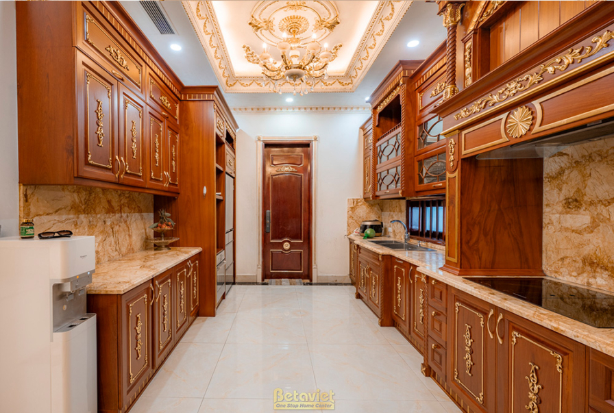 Phòng bếp với hệ tủ gỗ tự nhiên màu nâu đỏ, mặt đá granite. Bố cục tiện nghi, ánh sáng từ đèn downlight và đèn hắt. Không…