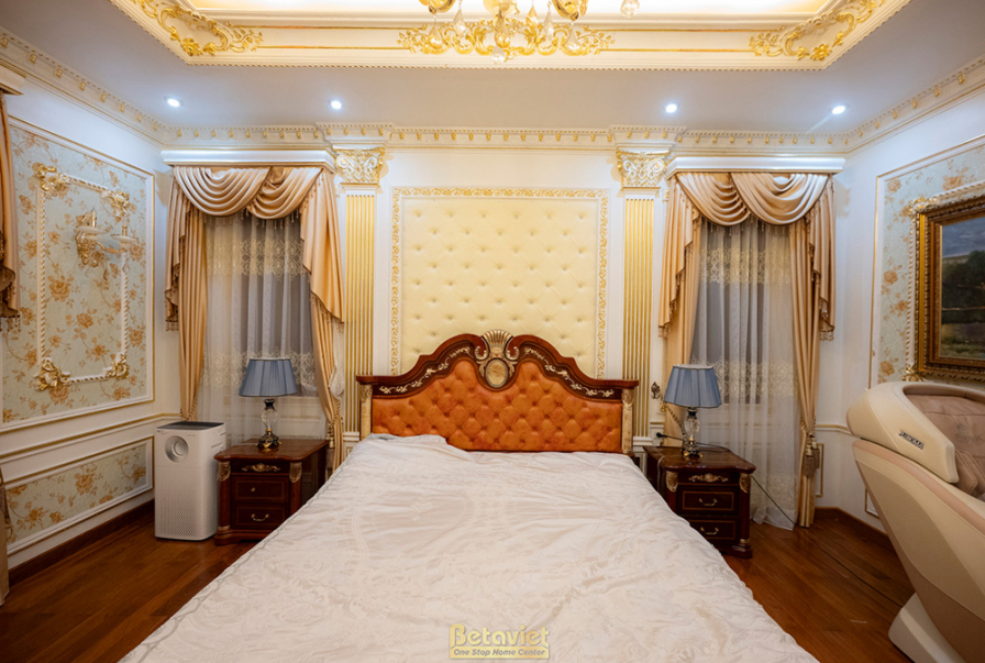 Phòng ngủ master với giường bọc nỉ màu kem, tủ đầu giường gỗ. Tường ốp phào chỉ và giấy dán tường họa tiết. Rèm cửa hai lớ…