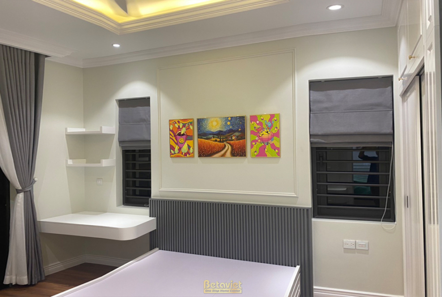 Phòng ngủ trẻ em với bàn học liền tủ, tường trang trí tranh màu sắc. Giường đơn, rèm cửa màu xám. Ánh sáng tự nhiên và đèn…
