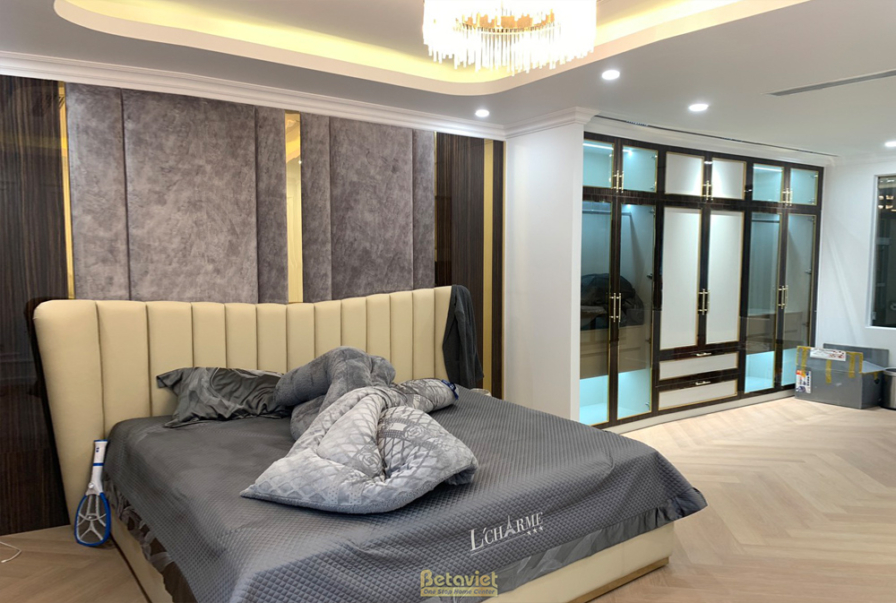 Phòng ngủ master với giường lớn bọc nỉ màu be, tường đầu giường ốp nỉ và gỗ viền kim loại vàng. Sàn gỗ xương cá, cửa kính…