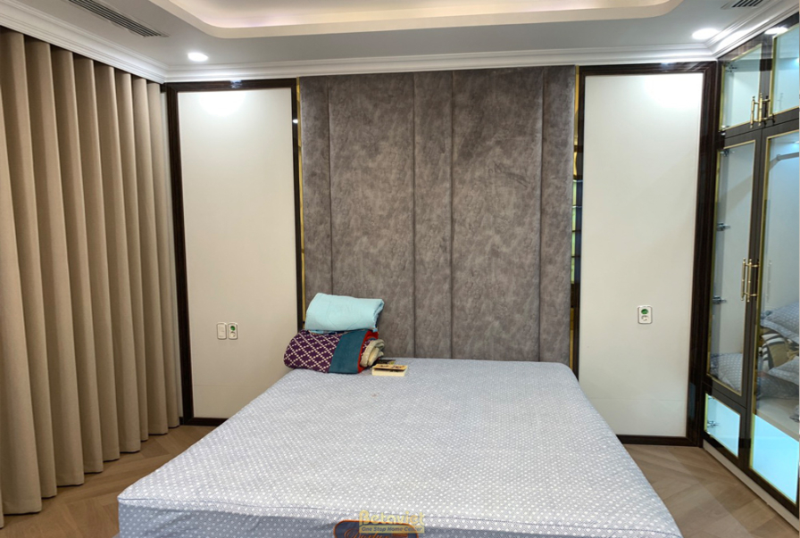 Phòng ngủ hiện đại với tường đầu giường ốp nỉ màu xám và gỗ, viền kim loại vàng. Giường lớn màu be, sàn gỗ xương cá. Ánh s…