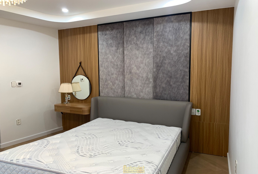 Phòng ngủ đơn giản với giường bọc nỉ màu xám, tủ đầu giường trắng. Tường ốp gỗ và đá vân xám, gương tròn trang trí, tạo kh…