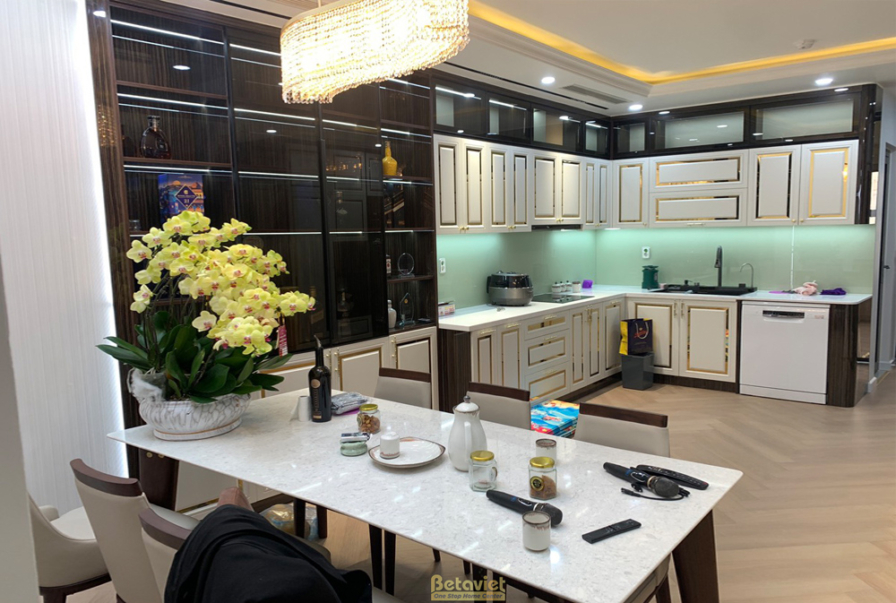 Khu vực bàn ăn và bếp mở, bàn ăn mặt đá trắng, ghế ăn bọc da. Hệ tủ bếp màu trắng kem viền vàng, đèn chùm pha lê, tạo khôn…