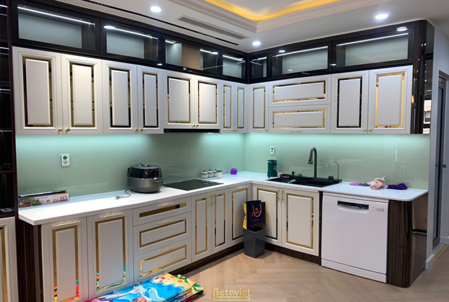 Góc bếp với hệ tủ trên dưới màu trắng kem viền vàng, mặt bếp đá trắng. Bồn rửa và bếp từ âm, không gian gọn gàng, sạch sẽ…