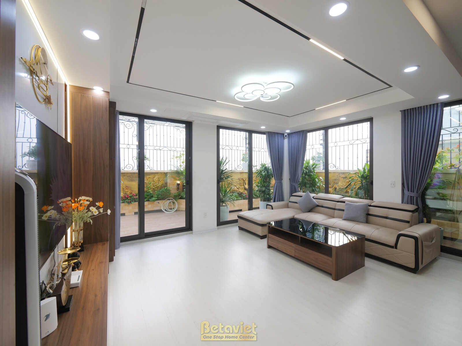 Phòng khách hiện đại nhìn từ góc sofa chữ L màu be, mở ra ban công xanh mát qua cửa kính lớn. Bố cục thoáng đãng, tràn ngậ…