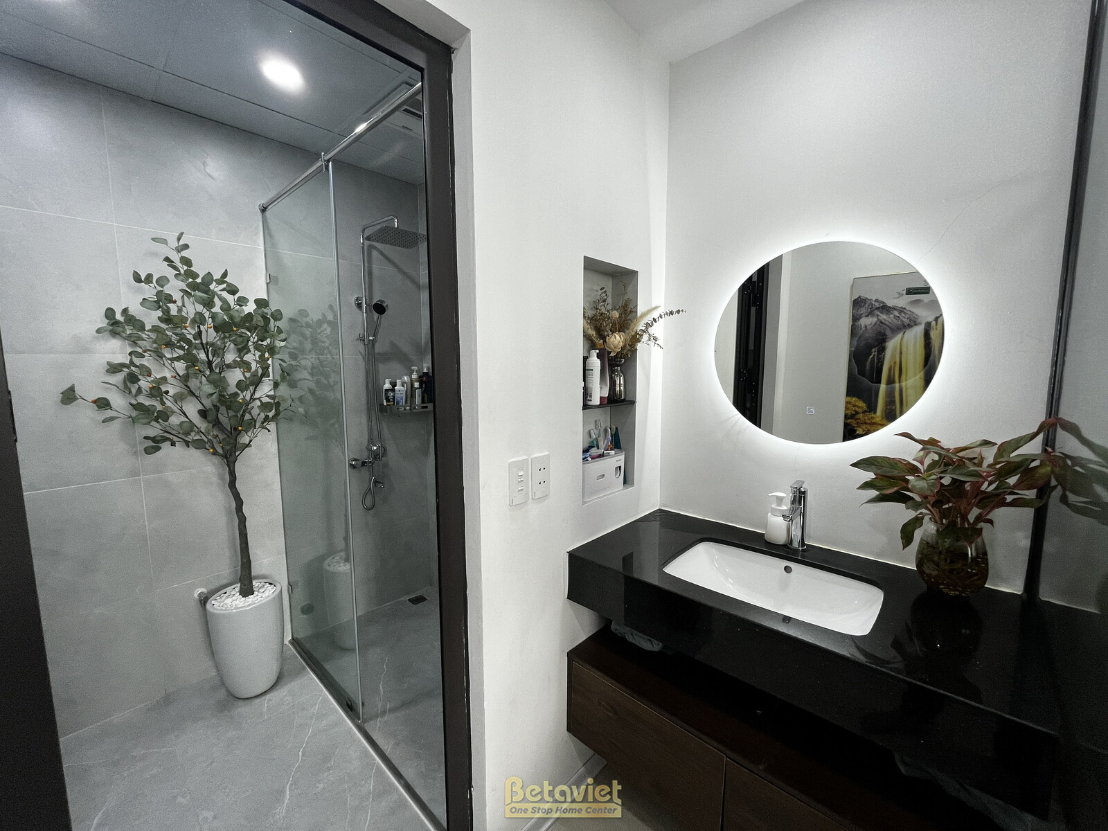 Không gian phòng tắm hiện đại với lavabo đặt trên mặt đá đen, gương tròn lớn có đèn LED hắt sáng. Tường ốp gạch màu xám nh…