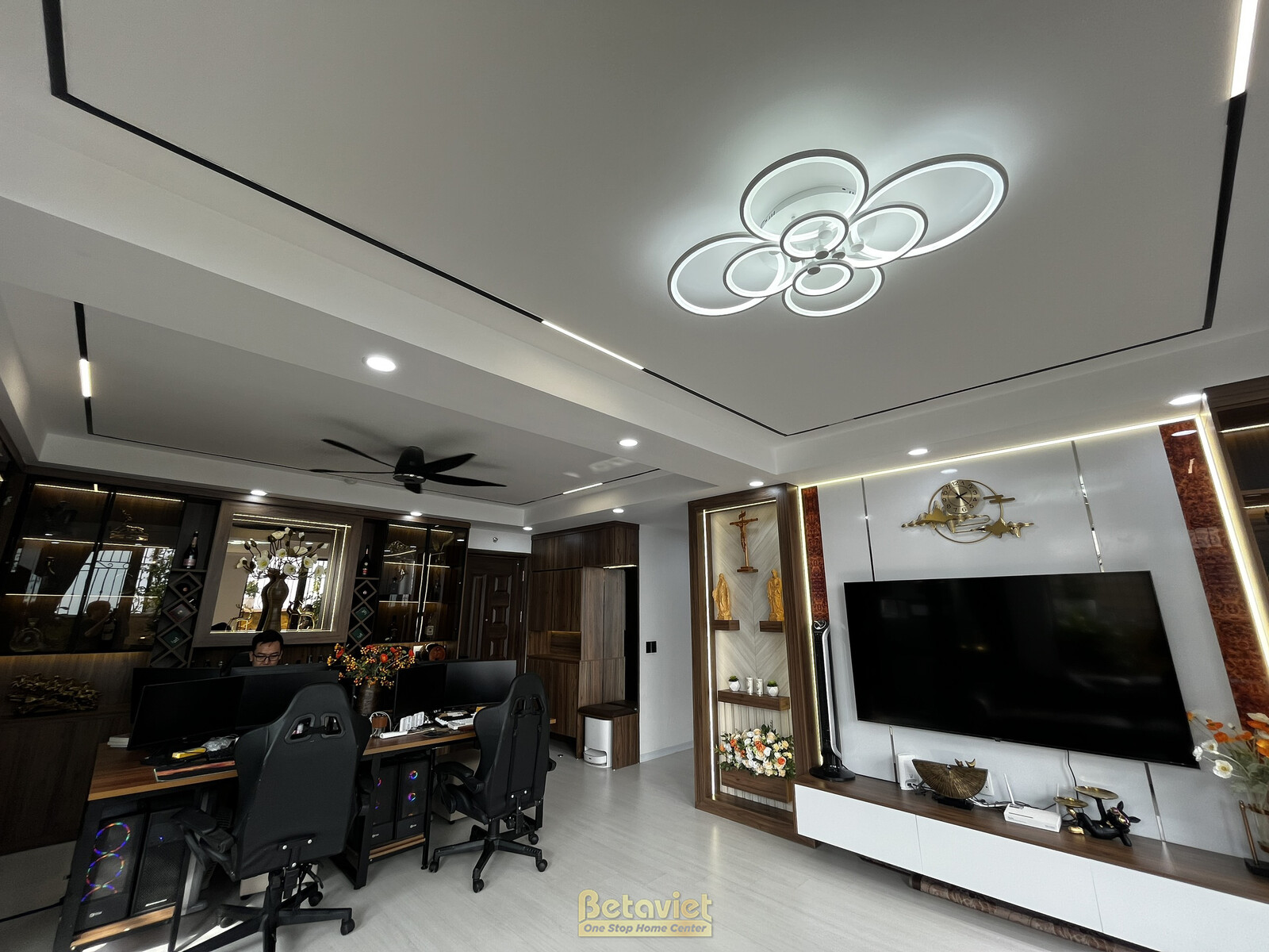 Cận cảnh góc phòng khách và bàn làm việc tích hợp. Bàn làm việc gỗ tối màu, ghế xoay đen. Trần giật cấp với đèn chùm hình…