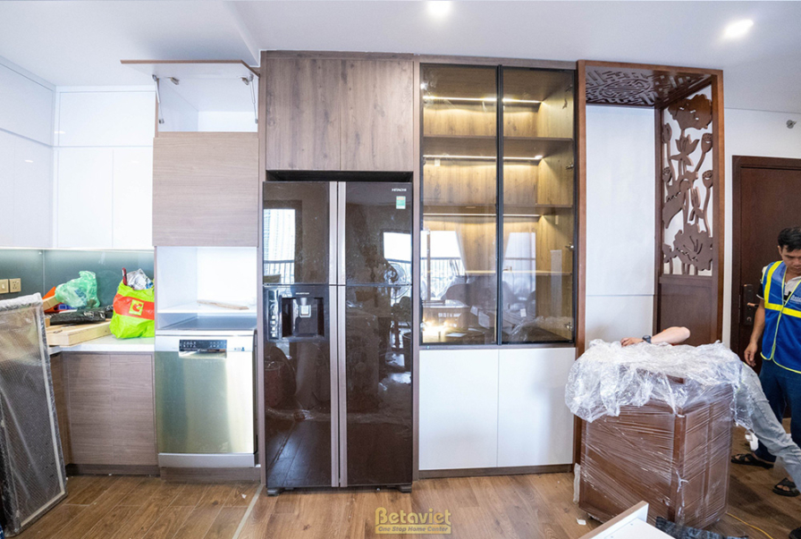 Khu vực bếp đang trong giai đoạn lắp đặt, với tủ lạnh âm tường và hệ tủ bếp kết hợp gỗ cùng kính. Một thợ đang kiểm tra ch…