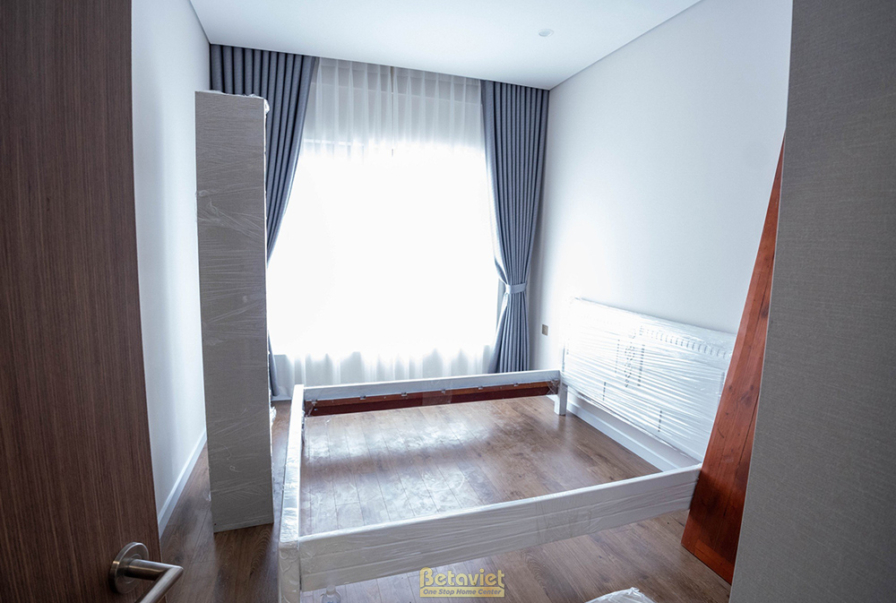 Cảnh thi công lắp đặt khung giường gỗ trong phòng ngủ, nhìn từ góc khác. Không gian đơn giản với sàn gỗ và tường trắng, ch…
