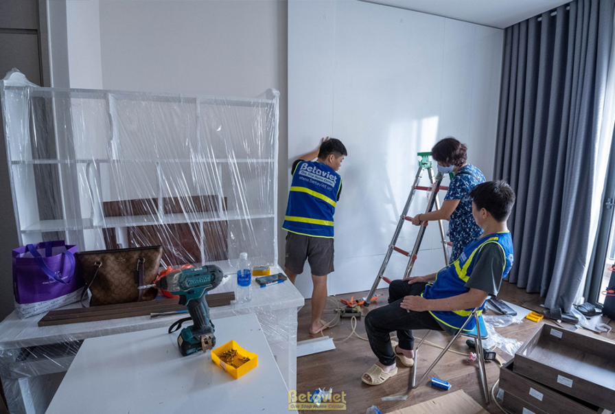Cảnh thi công lắp đặt kệ TV và tủ trang trí trong phòng khách. Các thợ đang tập trung vào việc cố định các tấm ván gỗ, đảm…