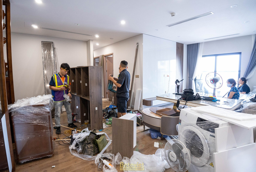Không gian phòng khách và bếp đang trong giai đoạn lắp đặt nội thất. Nhiều vật liệu, thùng carton và dụng cụ thi công được…