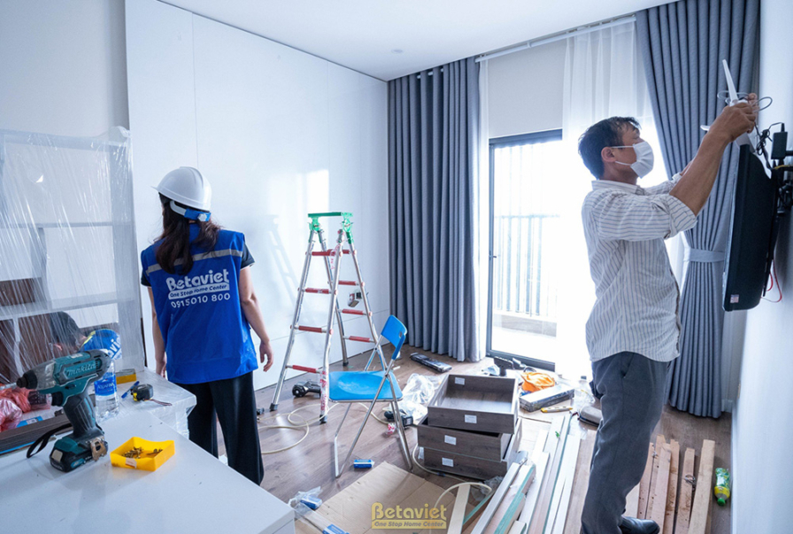 Cảnh thi công lắp đặt rèm cửa trong phòng ngủ, hai thợ đang kiểm tra và điều chỉnh. Không gian phòng ngủ với sàn gỗ, tường…