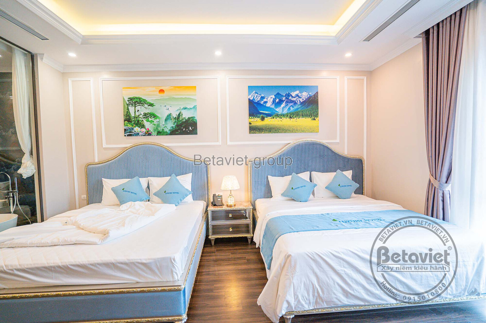 Phòng ngủ khách sạn với hai giường đơn, đầu giường bọc nệm và phào chỉ tường trang trí. Tông màu trắng chủ đạo kết hợp vàn…