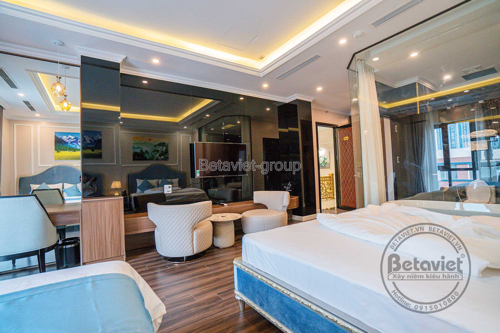 Phòng ngủ khách sạn với giường đôi lớn và khu vực tiếp khách nhỏ gồm bàn tròn và ghế bọc nệm. Vách kính trong suốt ngăn cá…